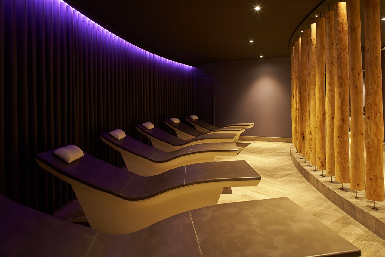 Curved lounge chairs sit empty, aligned in a row, under soft purple accent lights, within a dim spa relaxation room bordered by vertical wooden columns and dark ribbed wall panels.